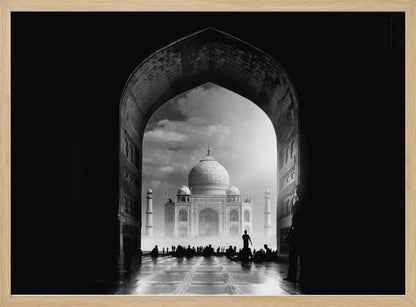 A dramatic black and white photograph of the Taj Mahal seen through a large, dark archway which frames the view. The iconic mausoleum is in the distance under a cloudy sky, and the silhouettes of many people are scattered on the wet, reflective ground in the foreground. Decor