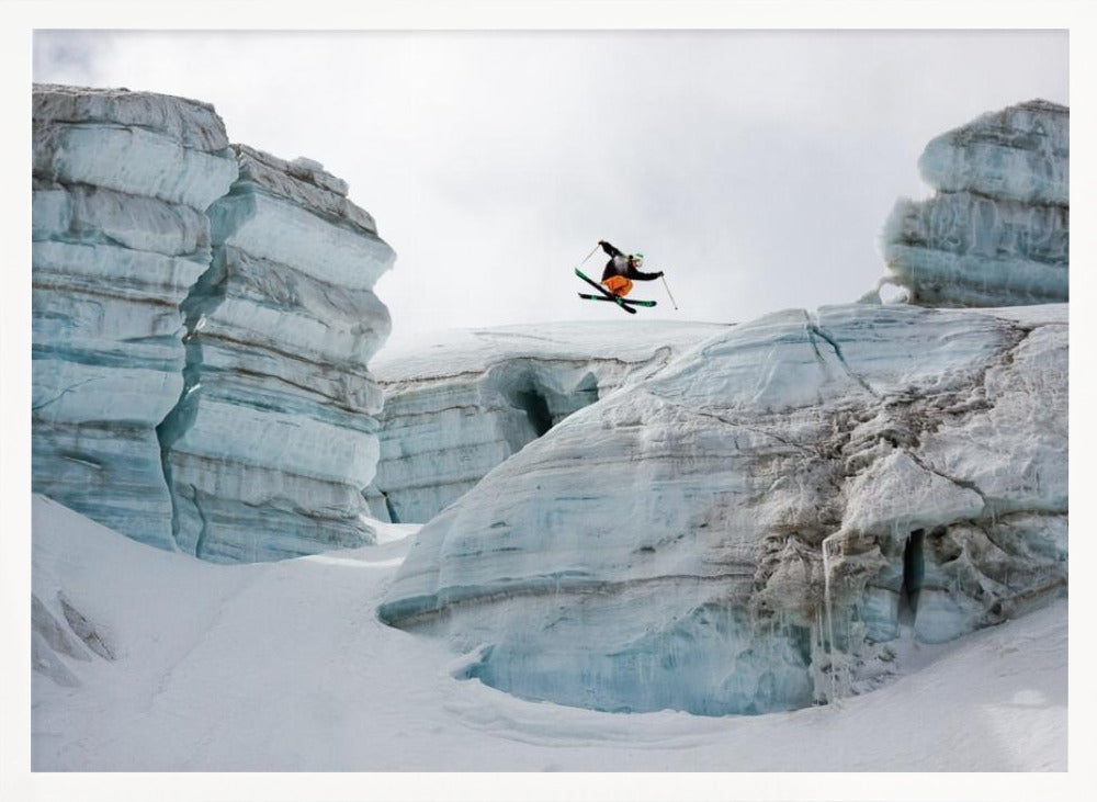 A framed photo of a skier in mid-air, jumping over a snowy crevasse between large, layered glacier formations under an overcast sky. The skier wears a black top and orange pants. Wall Art