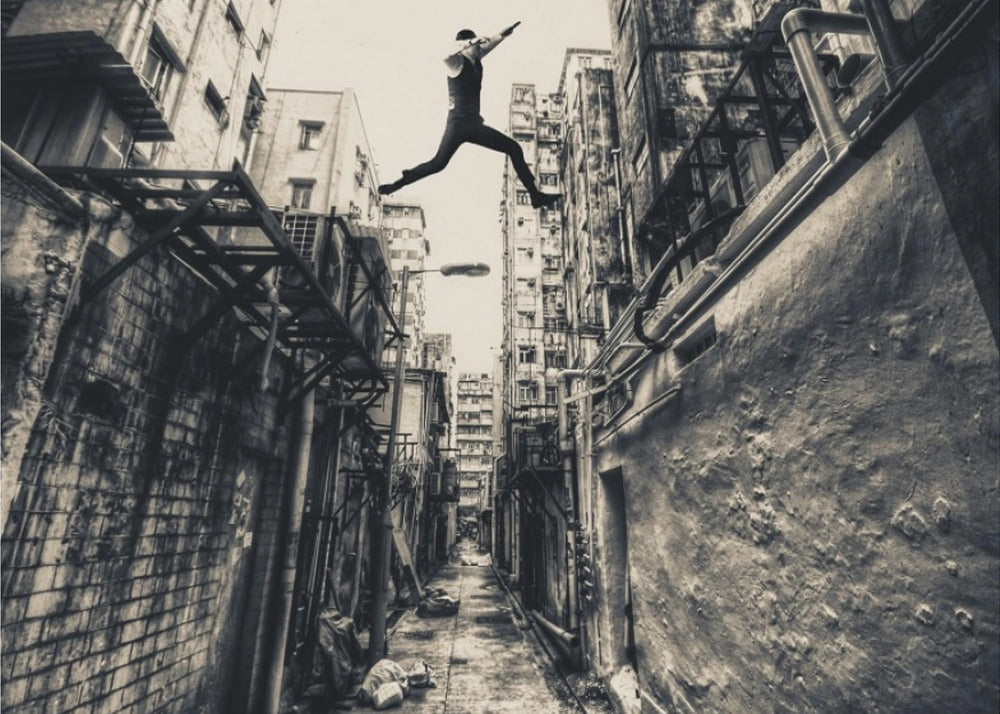 A dramatic black and white photograph of a man in mid-air, leaping athletically across a narrow, gritty urban alleyway. The low-angle shot looks up between tall, weathered buildings, emphasizing the height of the jump. The image is presented in a silver frame. Wall Art