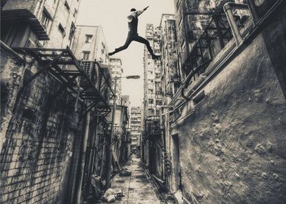 A dramatic black and white photograph of a man in mid-air, leaping athletically across a narrow, gritty urban alleyway. The low-angle shot looks up between tall, weathered buildings, emphasizing the height of the jump. The image is presented in a silver frame. Wall Art