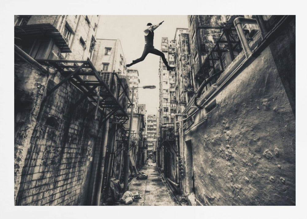 A dramatic black and white photograph of a man in mid-air, leaping athletically across a narrow, gritty urban alleyway. The low-angle shot looks up between tall, weathered buildings, emphasizing the height of the jump. The image is presented in a silver frame. Wall Art