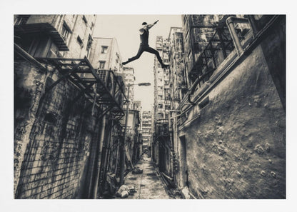 A dramatic black and white photograph of a man in mid-air, leaping athletically across a narrow, gritty urban alleyway. The low-angle shot looks up between tall, weathered buildings, emphasizing the height of the jump. The image is presented in a silver frame. Wall Art