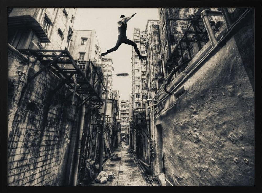 A dramatic black and white photograph of a man in mid-air, leaping athletically across a narrow, gritty urban alleyway. The low-angle shot looks up between tall, weathered buildings, emphasizing the height of the jump. The image is presented in a silver frame. Wall Art
