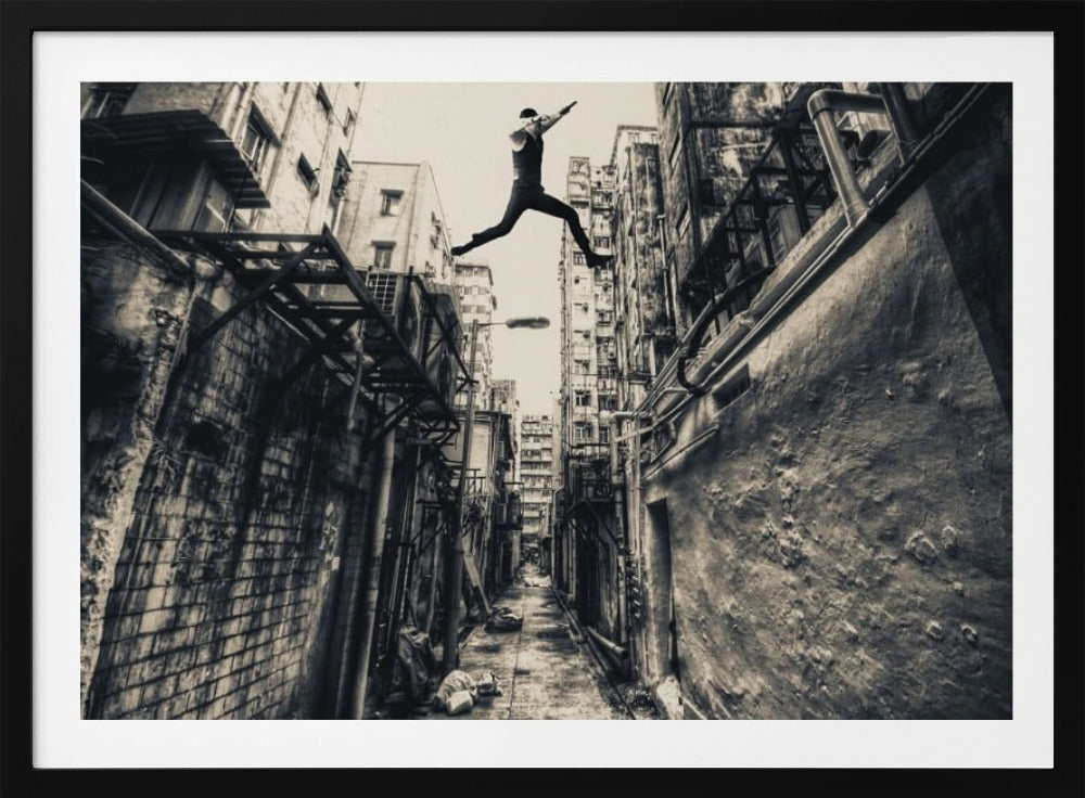 A dramatic black and white photograph of a man in mid-air, leaping athletically across a narrow, gritty urban alleyway. The low-angle shot looks up between tall, weathered buildings, emphasizing the height of the jump. The image is presented in a silver frame. Wall Art