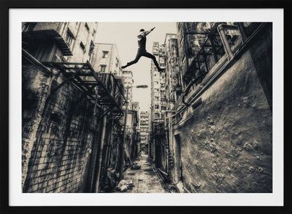A dramatic black and white photograph of a man in mid-air, leaping athletically across a narrow, gritty urban alleyway. The low-angle shot looks up between tall, weathered buildings, emphasizing the height of the jump. The image is presented in a silver frame. Wall Art
