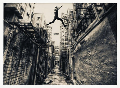 A dramatic black and white photograph of a man in mid-air, leaping athletically across a narrow, gritty urban alleyway. The low-angle shot looks up between tall, weathered buildings, emphasizing the height of the jump. The image is presented in a silver frame. Wall Art