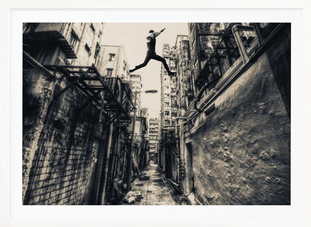 A dramatic black and white photograph of a man in mid-air, leaping athletically across a narrow, gritty urban alleyway. The low-angle shot looks up between tall, weathered buildings, emphasizing the height of the jump. The image is presented in a silver frame. Wall Art