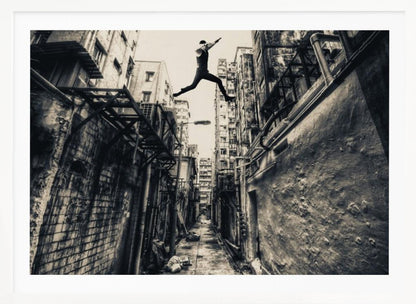 A dramatic black and white photograph of a man in mid-air, leaping athletically across a narrow, gritty urban alleyway. The low-angle shot looks up between tall, weathered buildings, emphasizing the height of the jump. The image is presented in a silver frame. Wall Art
