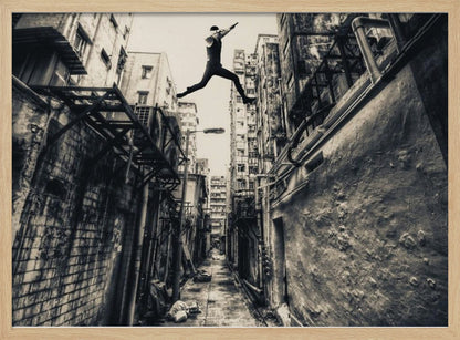 A dramatic black and white photograph of a man in mid-air, leaping athletically across a narrow, gritty urban alleyway. The low-angle shot looks up between tall, weathered buildings, emphasizing the height of the jump. The image is presented in a silver frame. Wall Art