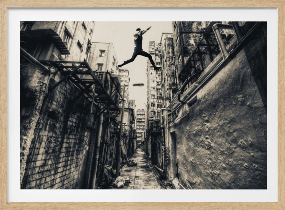 A dramatic black and white photograph of a man in mid-air, leaping athletically across a narrow, gritty urban alleyway. The low-angle shot looks up between tall, weathered buildings, emphasizing the height of the jump. The image is presented in a silver frame. Wall Art
