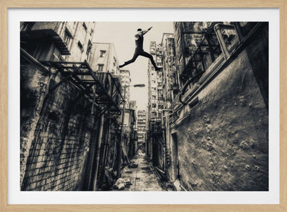 A dramatic black and white photograph of a man in mid-air, leaping athletically across a narrow, gritty urban alleyway. The low-angle shot looks up between tall, weathered buildings, emphasizing the height of the jump. The image is presented in a silver frame. Wall Art
