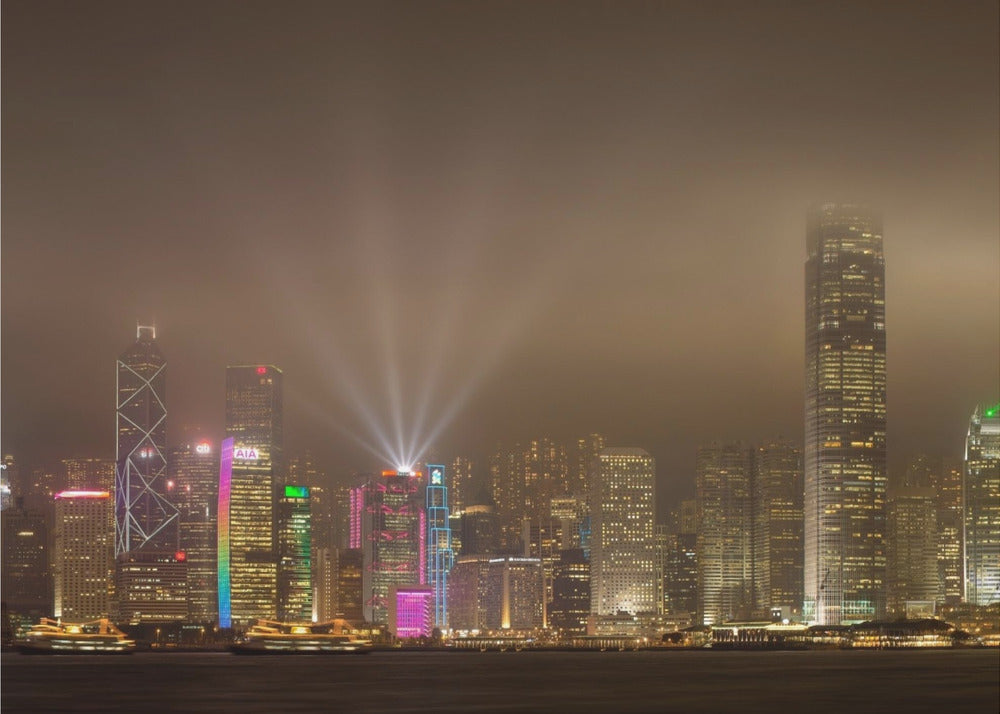 A framed photograph of the Hong Kong skyline at night, viewed from across the water. The skyscrapers are brilliantly lit with yellow, white, and colorful lights, and beams of light shoot up into the foggy, glowing sky. Decor