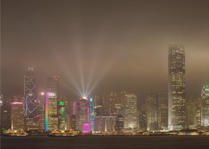 A framed photograph of the Hong Kong skyline at night, viewed from across the water. The skyscrapers are brilliantly lit with yellow, white, and colorful lights, and beams of light shoot up into the foggy, glowing sky. Decor