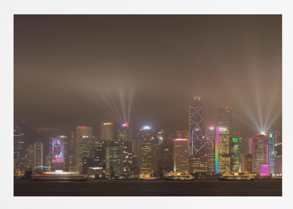 A framed photograph of the Hong Kong skyline at night, viewed from across the water. The skyscrapers are brilliantly lit with yellow, white, and colorful lights, and beams of light shoot up into the foggy, glowing sky. Decor