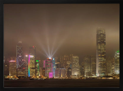 A framed photograph of the Hong Kong skyline at night, viewed from across the water. The skyscrapers are brilliantly lit with yellow, white, and colorful lights, and beams of light shoot up into the foggy, glowing sky. Decor