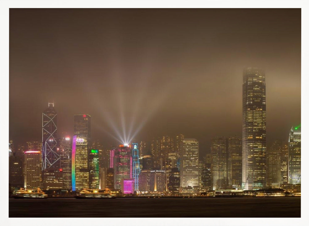 A framed photograph of the Hong Kong skyline at night, viewed from across the water. The skyscrapers are brilliantly lit with yellow, white, and colorful lights, and beams of light shoot up into the foggy, glowing sky. Decor