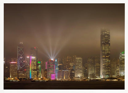 A framed photograph of the Hong Kong skyline at night, viewed from across the water. The skyscrapers are brilliantly lit with yellow, white, and colorful lights, and beams of light shoot up into the foggy, glowing sky. Decor