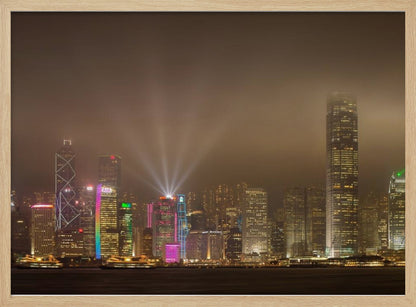 A framed photograph of the Hong Kong skyline at night, viewed from across the water. The skyscrapers are brilliantly lit with yellow, white, and colorful lights, and beams of light shoot up into the foggy, glowing sky. Decor