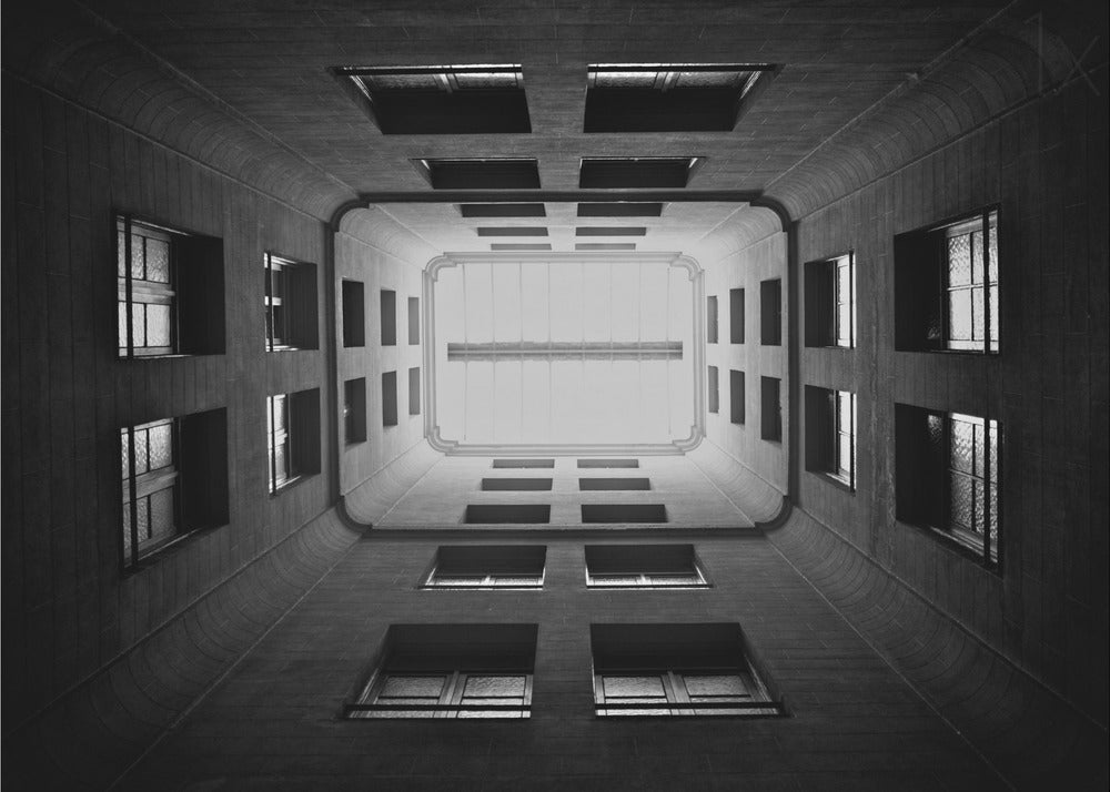 A symmetrical, black and white photograph looking directly up from the base of a building's interior courtyard. The dark walls rise up, lined with rows of windows, creating a powerful sense of perspective that draws the eye to the bright rectangle of the sky or a skylight at the top. The image is enclosed in a simple silver frame. Poster