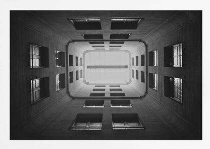 A symmetrical, black and white photograph looking directly up from the base of a building's interior courtyard. The dark walls rise up, lined with rows of windows, creating a powerful sense of perspective that draws the eye to the bright rectangle of the sky or a skylight at the top. The image is enclosed in a simple silver frame. Poster