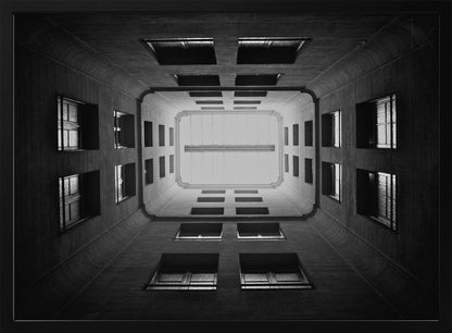 A symmetrical, black and white photograph looking directly up from the base of a building's interior courtyard. The dark walls rise up, lined with rows of windows, creating a powerful sense of perspective that draws the eye to the bright rectangle of the sky or a skylight at the top. The image is enclosed in a simple silver frame. Poster