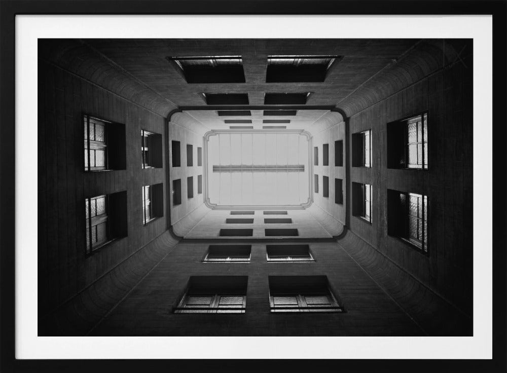 A symmetrical, black and white photograph looking directly up from the base of a building's interior courtyard. The dark walls rise up, lined with rows of windows, creating a powerful sense of perspective that draws the eye to the bright rectangle of the sky or a skylight at the top. The image is enclosed in a simple silver frame. Poster
