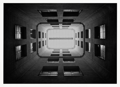 A symmetrical, black and white photograph looking directly up from the base of a building's interior courtyard. The dark walls rise up, lined with rows of windows, creating a powerful sense of perspective that draws the eye to the bright rectangle of the sky or a skylight at the top. The image is enclosed in a simple silver frame. Poster
