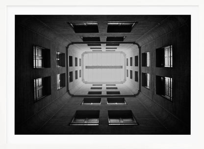 A symmetrical, black and white photograph looking directly up from the base of a building's interior courtyard. The dark walls rise up, lined with rows of windows, creating a powerful sense of perspective that draws the eye to the bright rectangle of the sky or a skylight at the top. The image is enclosed in a simple silver frame. Poster