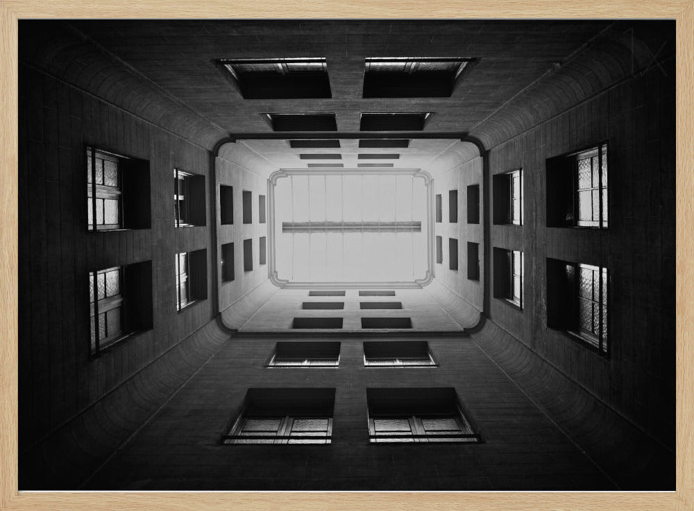 A symmetrical, black and white photograph looking directly up from the base of a building's interior courtyard. The dark walls rise up, lined with rows of windows, creating a powerful sense of perspective that draws the eye to the bright rectangle of the sky or a skylight at the top. The image is enclosed in a simple silver frame. Poster