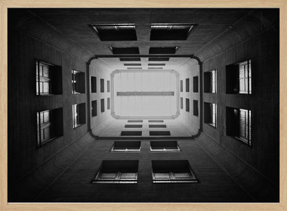 A symmetrical, black and white photograph looking directly up from the base of a building's interior courtyard. The dark walls rise up, lined with rows of windows, creating a powerful sense of perspective that draws the eye to the bright rectangle of the sky or a skylight at the top. The image is enclosed in a simple silver frame. Poster