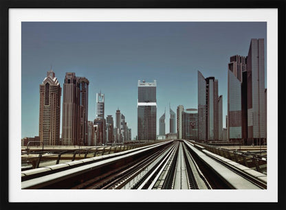 A view from an elevated train track looking down the rails towards the towering skyscrapers of a modern city skyline, under a clear, muted blue sky. Wall Art