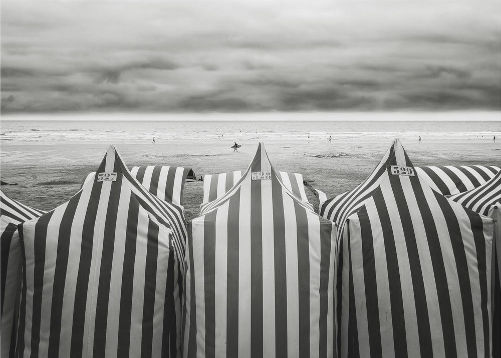 A framed, black and white photograph of striped beach tents on a cloudy day, with the ocean and distant surfers in the background. Print