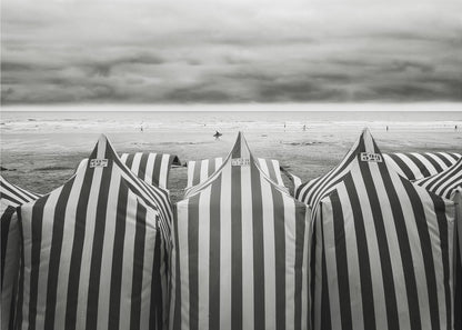 A framed, black and white photograph of striped beach tents on a cloudy day, with the ocean and distant surfers in the background. Print