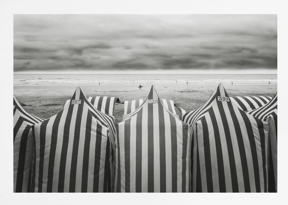 A framed, black and white photograph of striped beach tents on a cloudy day, with the ocean and distant surfers in the background. Print
