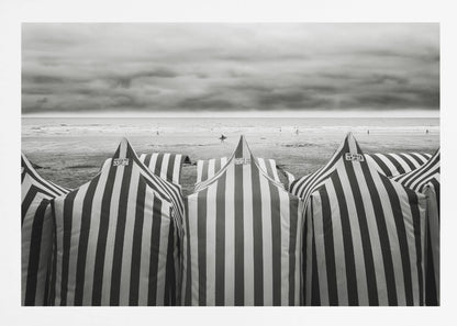 A framed, black and white photograph of striped beach tents on a cloudy day, with the ocean and distant surfers in the background. Print