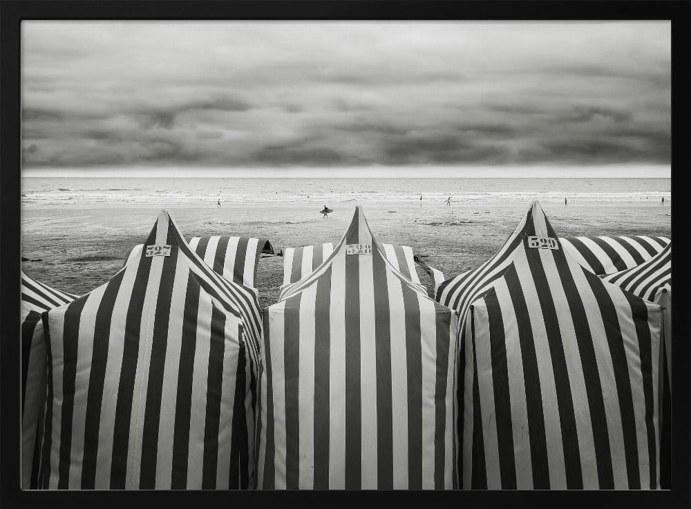A framed, black and white photograph of striped beach tents on a cloudy day, with the ocean and distant surfers in the background. Print