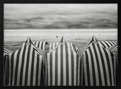 A framed, black and white photograph of striped beach tents on a cloudy day, with the ocean and distant surfers in the background. Print