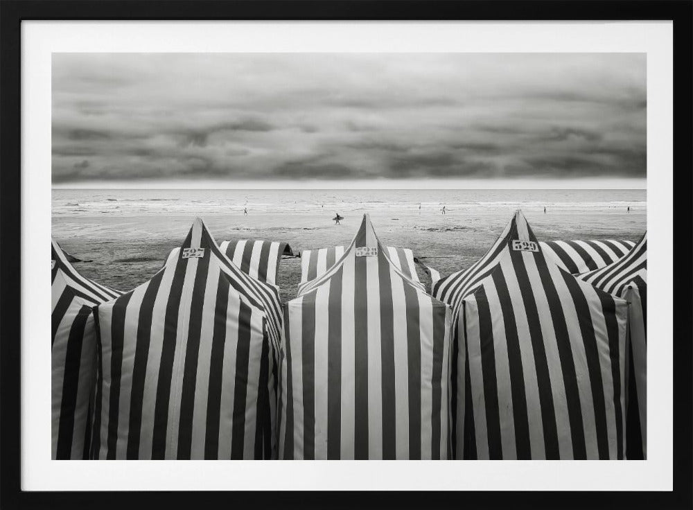 A framed, black and white photograph of striped beach tents on a cloudy day, with the ocean and distant surfers in the background. Print