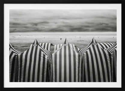 A framed, black and white photograph of striped beach tents on a cloudy day, with the ocean and distant surfers in the background. Print