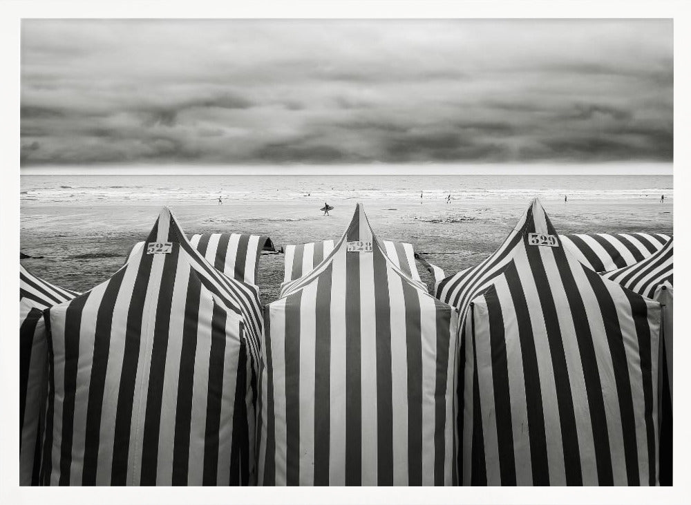 A framed, black and white photograph of striped beach tents on a cloudy day, with the ocean and distant surfers in the background. Print