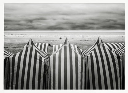 A framed, black and white photograph of striped beach tents on a cloudy day, with the ocean and distant surfers in the background. Print