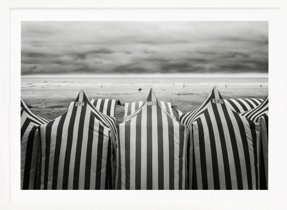 A framed, black and white photograph of striped beach tents on a cloudy day, with the ocean and distant surfers in the background. Print
