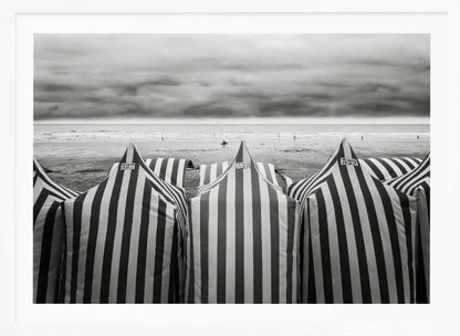 A framed, black and white photograph of striped beach tents on a cloudy day, with the ocean and distant surfers in the background. Print