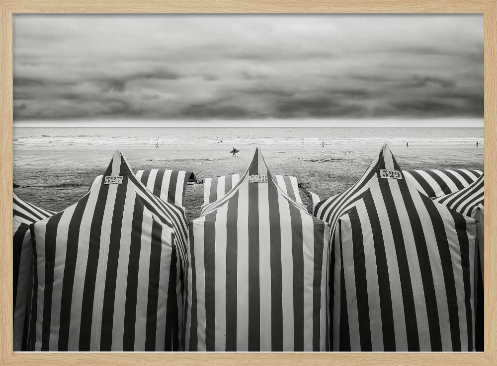 A framed, black and white photograph of striped beach tents on a cloudy day, with the ocean and distant surfers in the background. Print