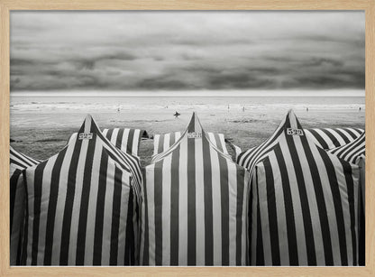 A framed, black and white photograph of striped beach tents on a cloudy day, with the ocean and distant surfers in the background. Print