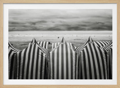 A framed, black and white photograph of striped beach tents on a cloudy day, with the ocean and distant surfers in the background. Print