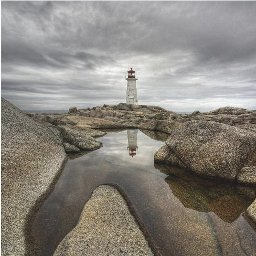 A framed photograph of a white lighthouse with a red top standing on a rocky shore under a dramatic, cloudy sky. In the foreground, a tide pool perfectly reflects the lighthouse. Artwork