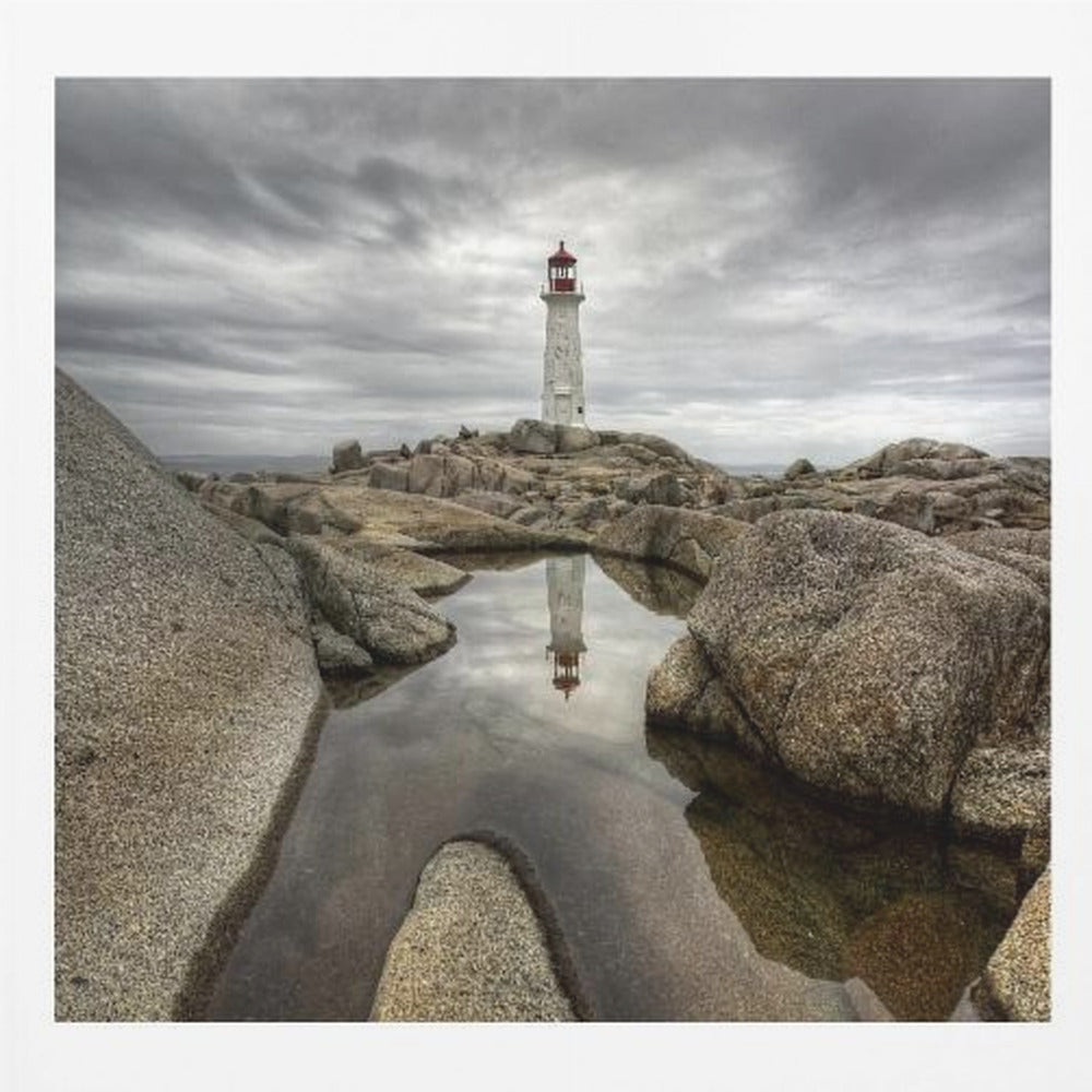 A framed photograph of a white lighthouse with a red top standing on a rocky shore under a dramatic, cloudy sky. In the foreground, a tide pool perfectly reflects the lighthouse. Artwork