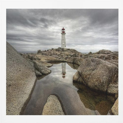 A framed photograph of a white lighthouse with a red top standing on a rocky shore under a dramatic, cloudy sky. In the foreground, a tide pool perfectly reflects the lighthouse. Artwork