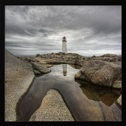 A framed photograph of a white lighthouse with a red top standing on a rocky shore under a dramatic, cloudy sky. In the foreground, a tide pool perfectly reflects the lighthouse. Artwork